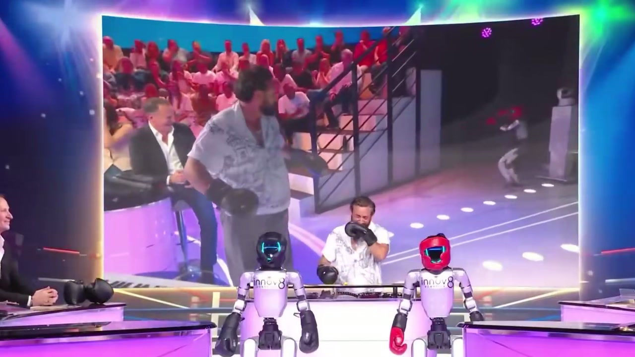 The robots at the studio desk as the big screen replays the fight (June 2026)